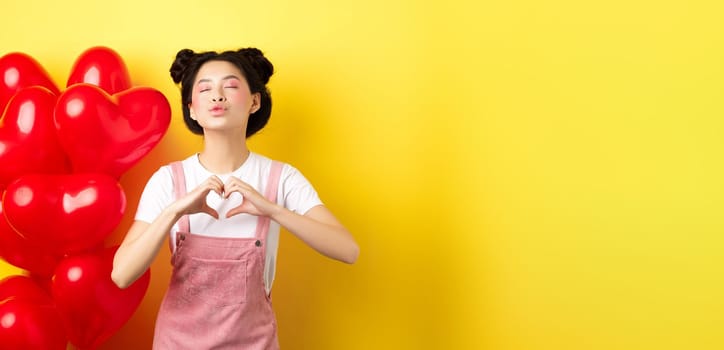 Romantic girl close eyes and pucker lips for kiss, showing I love you heart gesture, standing near cute red balloons, kissing lover over yellow background.
