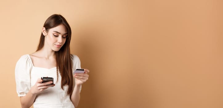 Smiling young woman paying online, looking at credit card and making purchase on mobile phone, shopping in internet, standing on beige background.
