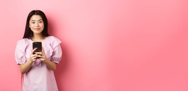 Online shopping. Cute asian girl smiling, holding mobile phone with happy face, standing in dress on pink background.