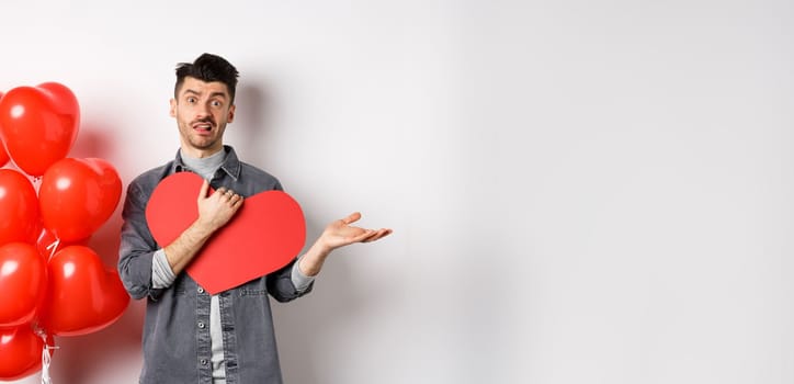 Confused man hugging big Valentine card and shrugging, feeling careless about romantic holiday, standing skeptical on white background.