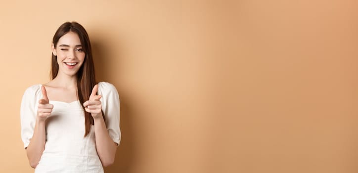 We need you. Smiling young woman pointing fingers at camera to beckon or invite people, standing on beige background.