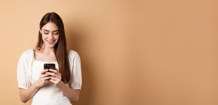 Young woman texting message on mobile phone, smiling and reading smartphone screen, standing on beige background.