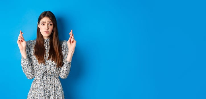 Nervous young woman in dress cross fingers and biting lip worried, praying or making wish, standing on blue background.