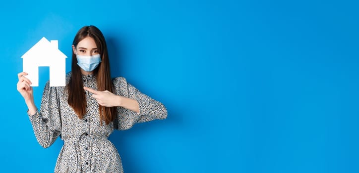 Real estate, covid-19 and pandemic concept. Smiling female model in medical mask pointing at paper house cutout, standing on blue background.