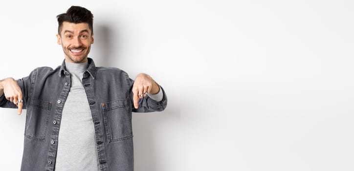 Happy caucasian man with moustache pointing fingers down, smiling and showing special deal, inviting to look here, standing on white background.