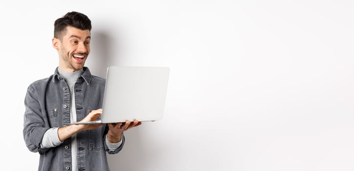 Man watching funny video on laptop, laughing at computer screen and smiling joyful, standing on white background.
