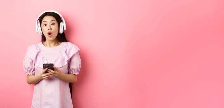 Excited asian woman in wireless headphones say wow, holding mobile phone and looking amused at camera, standing against pink background.
