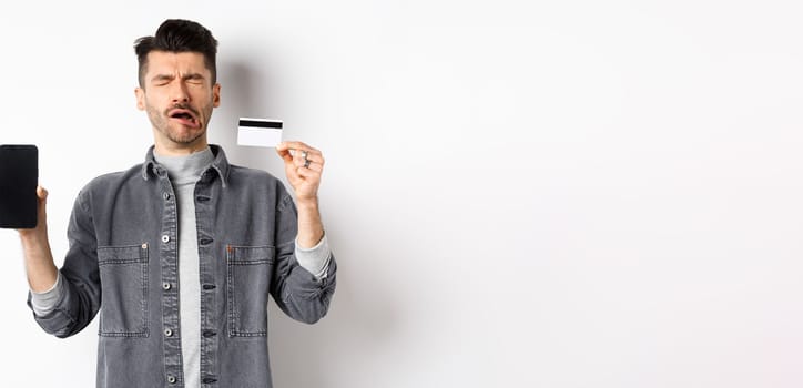 Sad crying man show empty mobile phone screen and plastic credit card, standing miserable on white background.