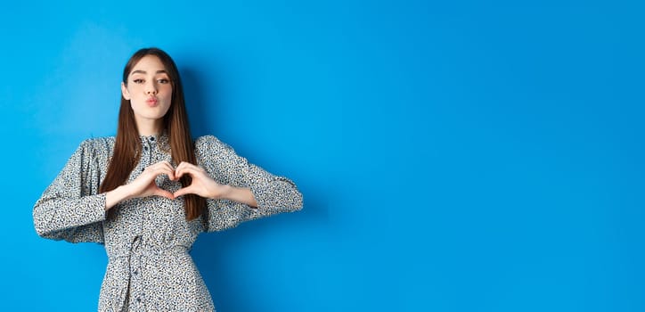 Valentines day. Romantic girl pucker lips for kiss, showing heart gesture, say I love you and look at camera, standing in dress on blue background.