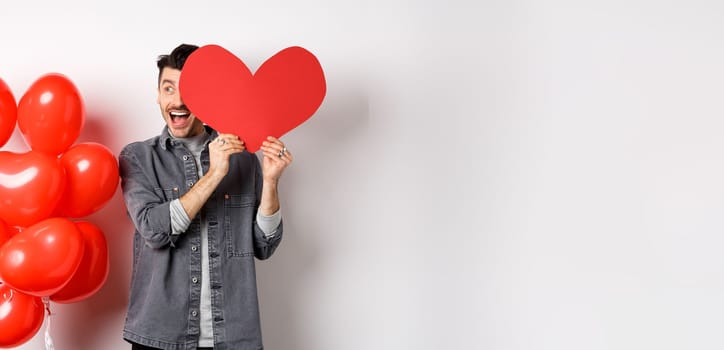 Romantic cheerful man cover half of face with Valentine heart cutout and smiling amazed, celebrating love holiday, standing on white background.