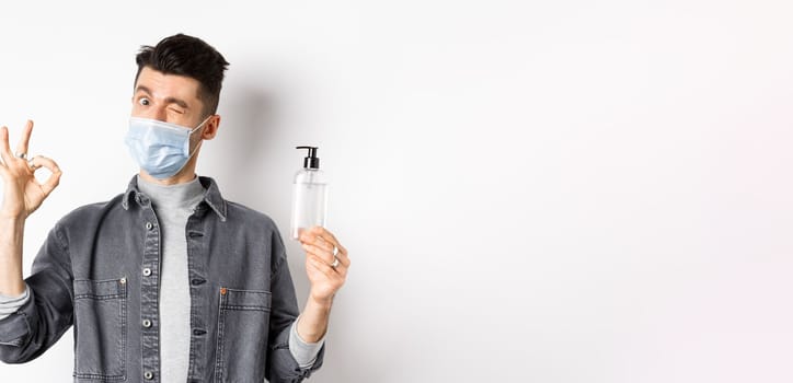 Healthy people and covid-19 concept. Excited man in medical mask holding bottle of good hand sanitizer, show okay sign, recommend antiseptic, standing against white background.