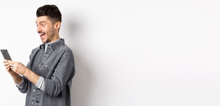 Profile portrait of excited handsome man reading message on phone, looking happy at smartphone screen, standing on white background.