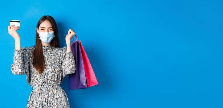 Covid-19, pandemic and lifestyle concept. Pretty female model in medical mask, rejoicing over shopping discounts, holding plastic credit card and bags, standing happy on blue background.