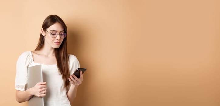 Young girl in glasses chatting on mobile phone and holding laptop, working, standing on beige background.