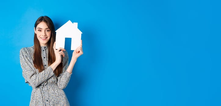 Real estate, realtors and insurance concept. Beautiful lady in dress showing paper house cutout, suggest property for sale, standing on blue background.