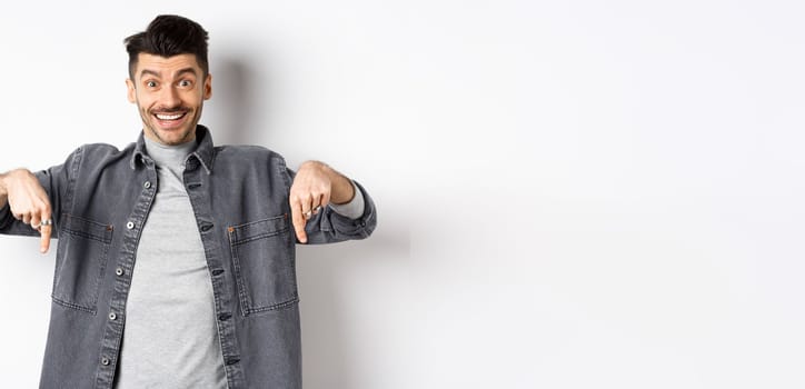 Excited smiling male model pointing fingers down, showing logo with happy face, demonstrate cool promo, standing on white background.