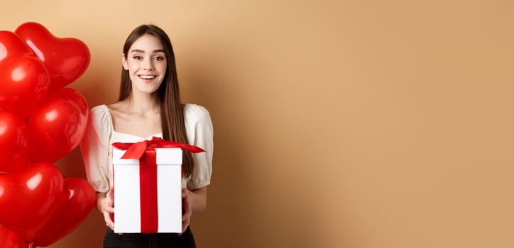 Romantic girl standing near heart balloons and holding surprise gift on Valentines day, smiling at camera happy, standing on beige background.