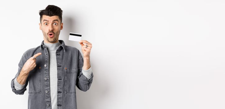 Wow look here. Excited guy pointing at plastic credit card and look amazed with awesome deal, standing against white background.