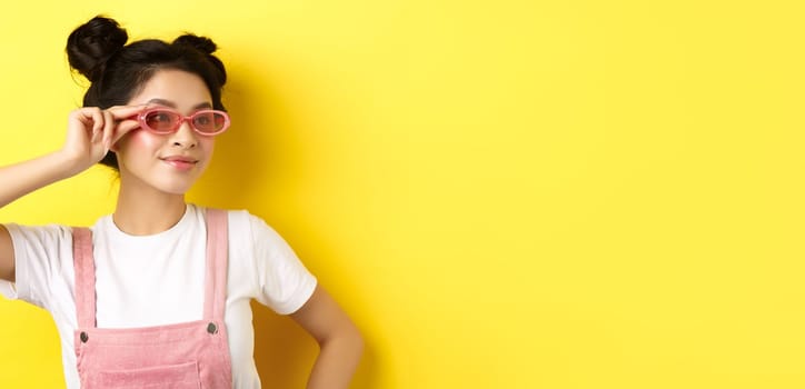 Beautiful asian girl put on stylish sunglasses and smiling, wearing trendy summer outfit, standing on yellow background.