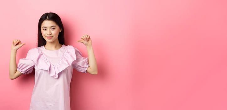 Confident asian girl smiling, pointing at herself, self-promoting personal achievements, standing in dress on pink background.