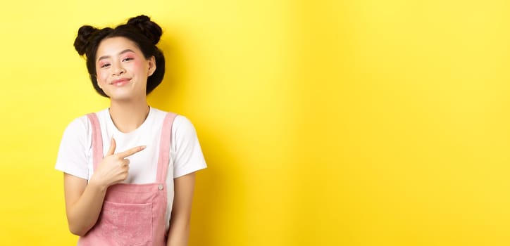 Stylish asian teen girl with romantic makeup, smiling cute and pointing finger right at logo, standing against yellow background.