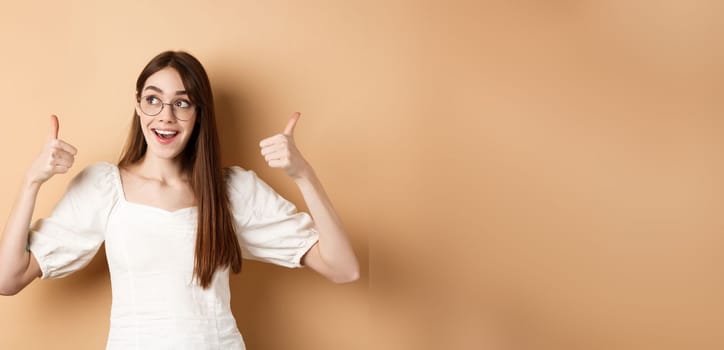 Happy girl in glasses show thumbs up, looking aside at good logo, standing satisfied on beige background.