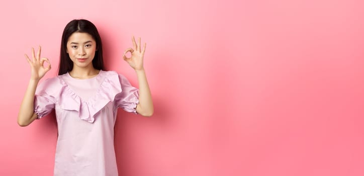 Smiling asian woman showing okay signs and looking confident, assure all good, praise good work, nice choice gesture, standing on pink background.