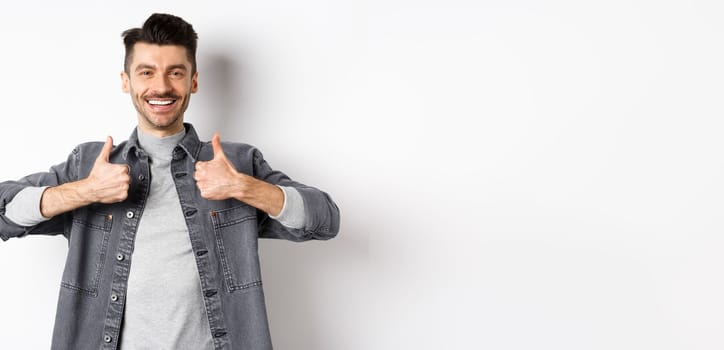 Handsome smiling man showing thumbs-up in approval, praise good work, satisfied with nice choice, recommending something cool, standing on white background.