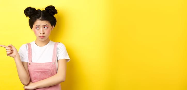 Upset frowning teenage girl sulking, pointing fingers left with jealous unfair face, standing disappointed on yellow background.