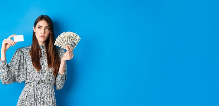 Shopping. Thoughtful girl looking up and biting lip, counting in mind or thinking, holding plastic credit card with dollar bills, standing on blue background.