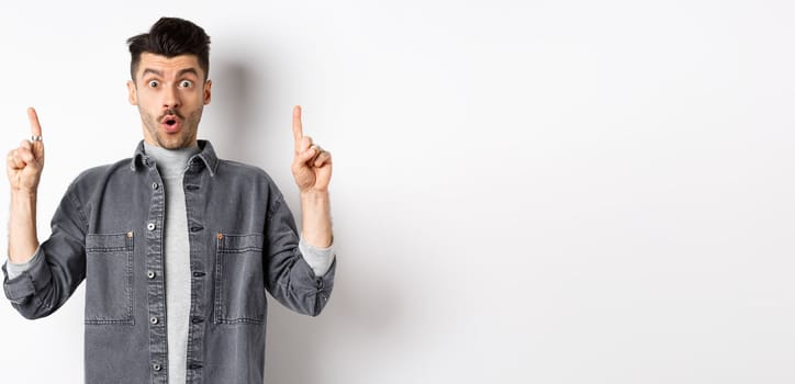 Excited man checking out promo offer, gasping amazed and pointing fingers up, making cool announcement, standing on white background.