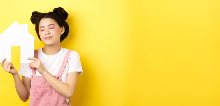 Real estate and family concept. Dreamy smiling asian woman with bright makeup, showing paper house cutout with closed eyes, daydreaming about buying property, yellow background.