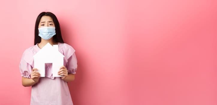 Real estate and pandemic concept. Begging asian girl showing paper house cutout, wearing medical mask, asking for apartment, standing against pink background.