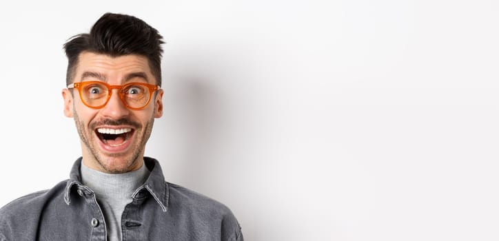 Excited funny man in glasses smiling, look with amazement and joy at awesome promo, standing happy against white background.