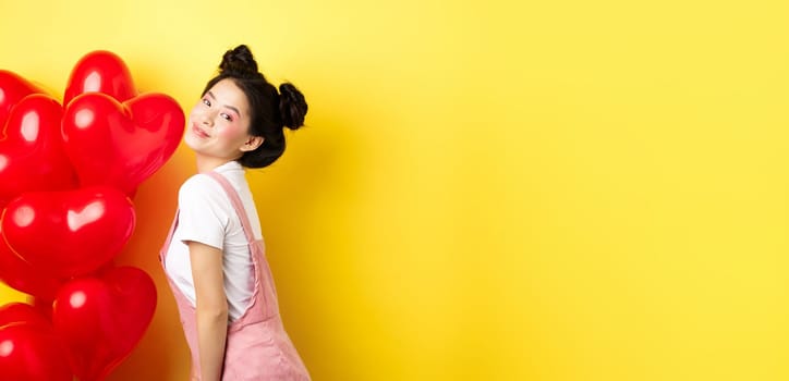 Valentines day and relationship concept. Fashionable asian woman posing near red hearts balloons, surprise for lover on special date, yellow background.