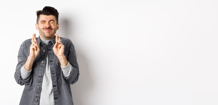 Hopeful young man standing nervous with closed eyes and fingers crossed, making wish, begging god for dream come true, waiting news, standing on white background.