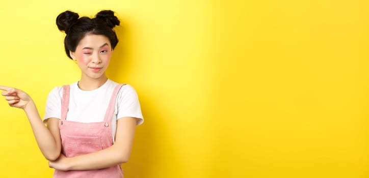 Glamour asian girl with bright makeup, pointing finger left and winking at camera, hint on good promo deal, standing on yellow background.