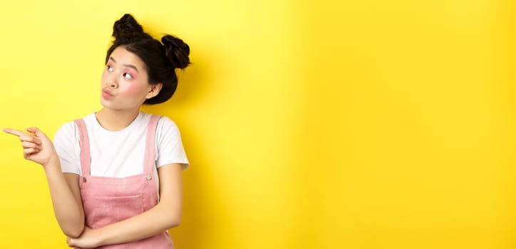 Pensive asian woman with beauty makeup, pointing and looking left with curious face, interested in advertisement, standing on yellow background.