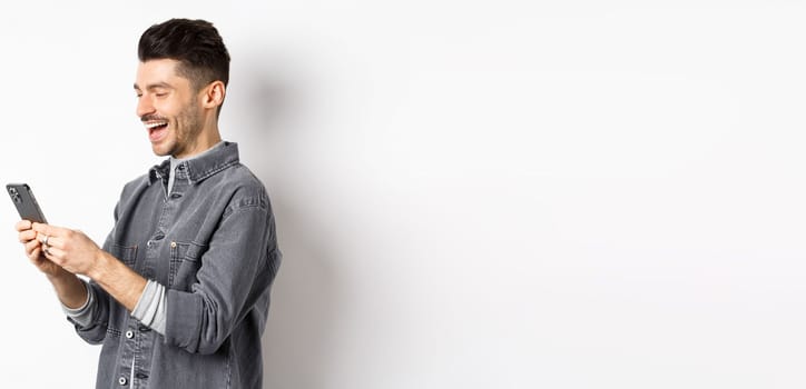 Profile of handsome man chatting on phone, laughing and reading smartphone screen, standing on white background.