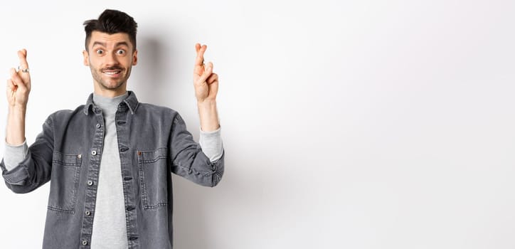 Hope dream come true. Positive and handsome guy making wish with fingers crossed, looking hopeful and smiling at camera, waiting results, standing on white background.