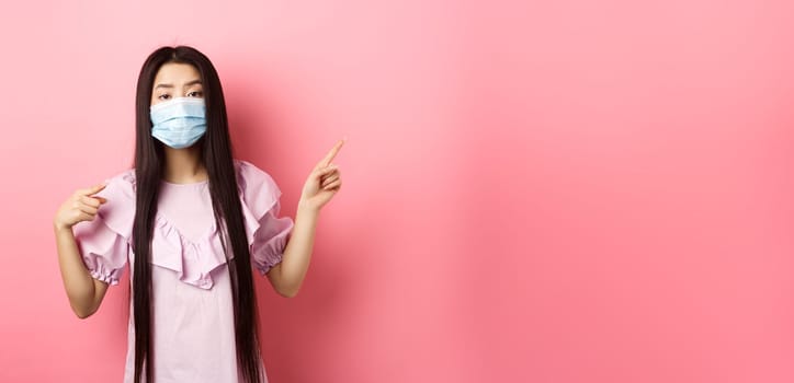 Healthy people and covid-19 pandemic concept. Bored asian woman in medical mask pointing right, showing logo, standing unamused on pink background.