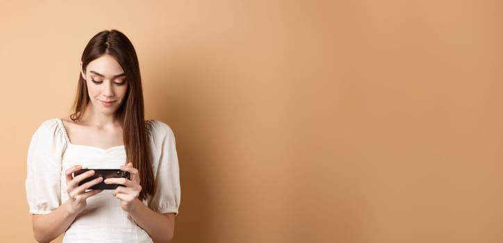 Pretty girl watching videos on smartphone, holding mobile phone horizontally and looking at screen, standing on beige background.