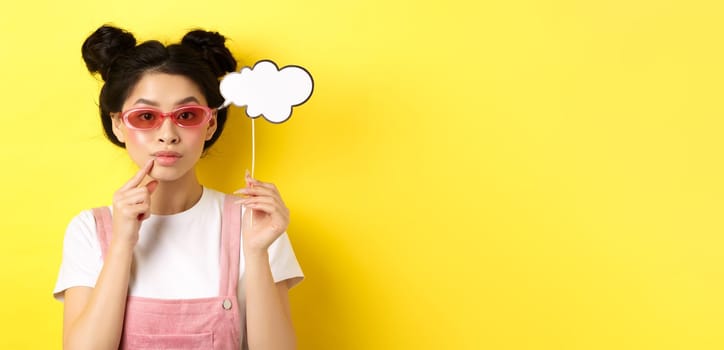 Summer and fashion concept. Stylish asian girl in sunglasses holding comment cloud on stick and looking thoughtful, thinking or saying something, standing on yellow background.