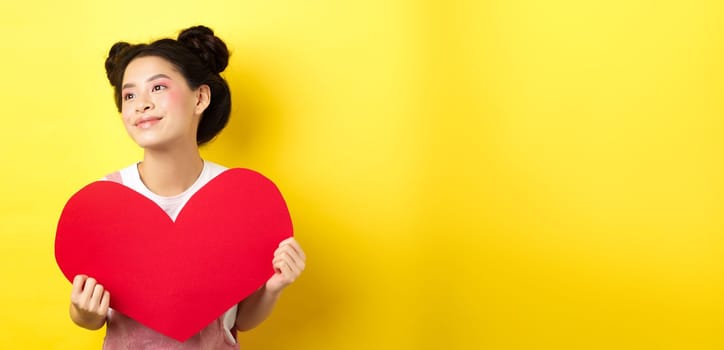 Asian girl in love, looking dreamy left and smiling, showing Valentines red heart cutout, imaging romantic date, standing on yellow background.