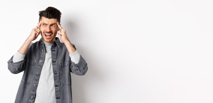 Distressed young man touching head and shouting, looking frustrated aside, standing on white background.