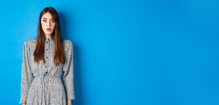 Shocked caucasian woman with long hair, wearing dress, gasping and standing in stupid against blue background, stare with disbelief.