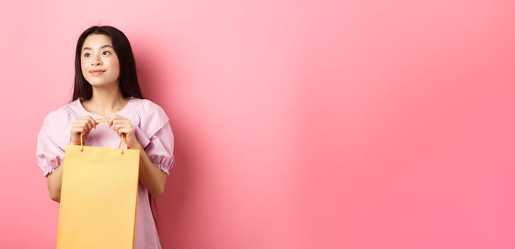 Beautiful asian girl dreaming and holding shopping bag, standing on pink background.