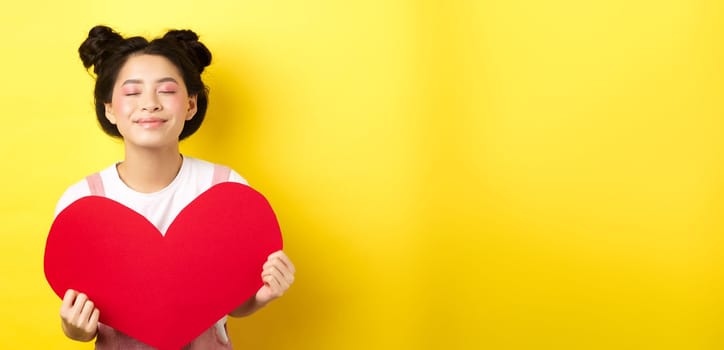 Cute teenage asian girl dreaming of true love, close eyes and showing big red heart cutout, waiting for soulmate on Valentines day, standing with romantic makeup on yellow background.