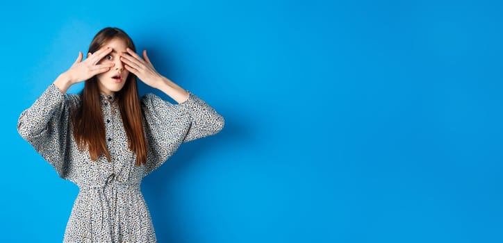 Intrigued young girl peeking through fingers with excitement, seeing interesting thing, standing on blue background.