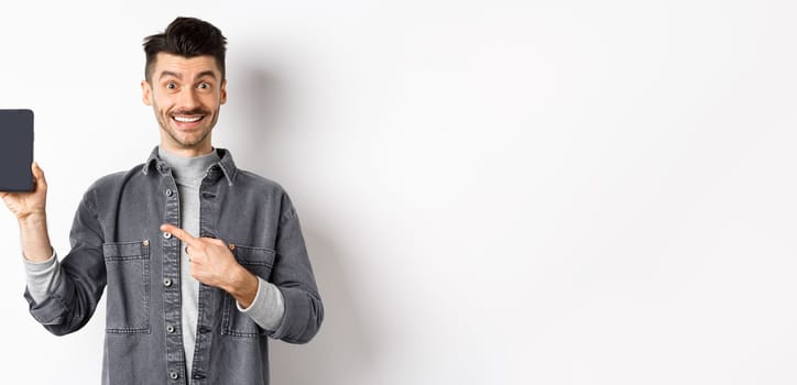 Handsome smiling man pointing finger at smartphone screen, showing mobile phone display with happy face, standing against white background.